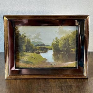 May include: A rectangular picture frame with a polished metallic gold finish containing a landscape print. The artwork depicts a calm river flowing through a lush green meadow with dense trees under a bright sky, reflecting light on the water surface.