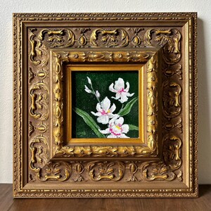 May include: A framed painting of white orchids with pink accents against a dark green background. The painting is in a gold-colored ornate frame with detailed carvings. The artwork is square and appears to be an oil painting.