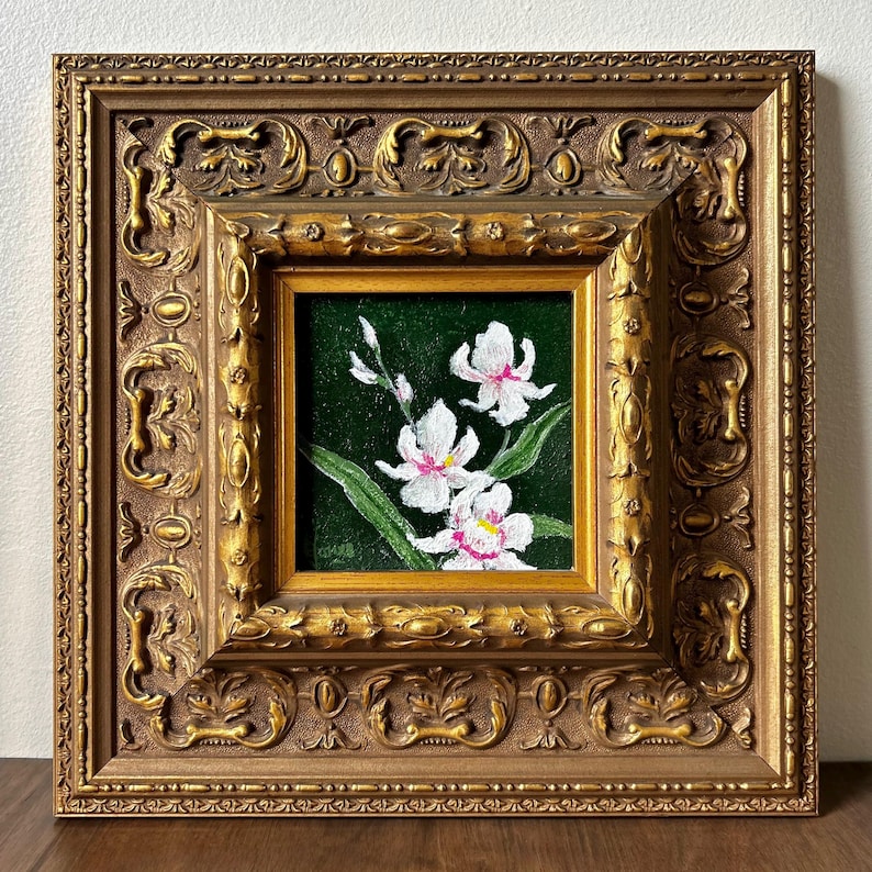 May include: A framed painting of white orchids with pink accents against a dark green background. The painting is in a gold-colored ornate frame with detailed carvings. The artwork is square and appears to be an oil painting.