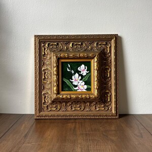 May include: A framed floral painting featuring white orchids with pink accents against a dark green background. The painting is set within a detailed, ornate gold-colored frame, showcasing a classic aesthetic. The artwork is displayed on a wooden surface.