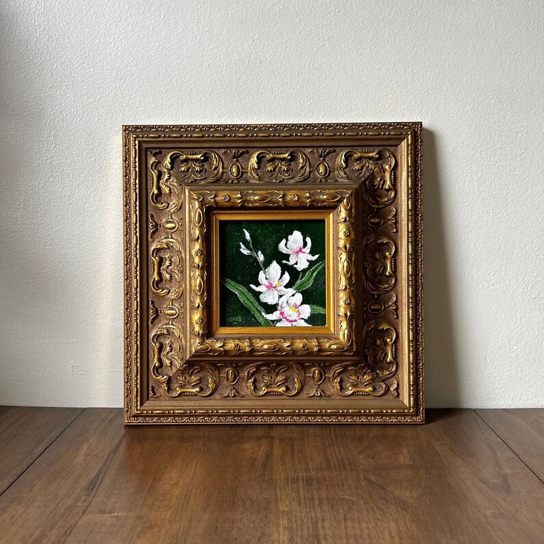 May include: A framed floral painting featuring white orchids with pink accents against a dark green background. The painting is set within a detailed, ornate gold-colored frame, showcasing a classic aesthetic. The artwork is displayed on a wooden surface.