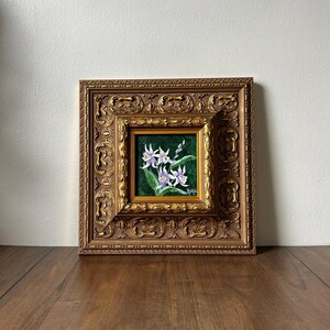 May include: A framed floral painting featuring white and purple flowers with green leaves against a dark green background. The painting is set within a detailed, ornate gold-colored frame, creating a classic aesthetic.