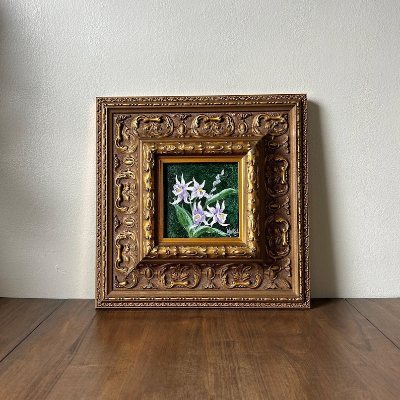 May include: A framed floral painting featuring white and purple flowers with green leaves against a dark green background. The painting is set within a detailed, ornate gold-colored frame, creating a classic aesthetic.