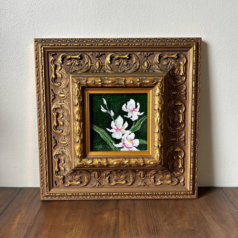 May include: A framed painting of white orchids with pink accents against a dark green background. The painting is set within a smaller gold frame, which is then set within a larger, ornate gold frame. The frames are resting on a wooden surface.