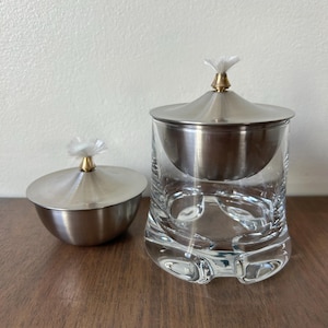 May include: Two glass and metal tea infusers. One is a small bowl with a lid and a white flower-shaped top. The other is a larger glass with a metal infuser and lid. The lids have a gold-colored base and a white flower-shaped top.
