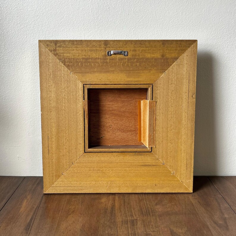May include: Back view of a square wooden frame with a recessed center. The frame is a light brown color with a natural wood grain. A metal hanger is attached to the top. The center is a darker brown color.