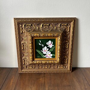 May include: A framed floral painting featuring white orchids with pink accents against a dark green background. The painting is set within a smaller gold frame, which is then encased in a larger, ornate gold frame. The artwork is displayed on a wooden surface.