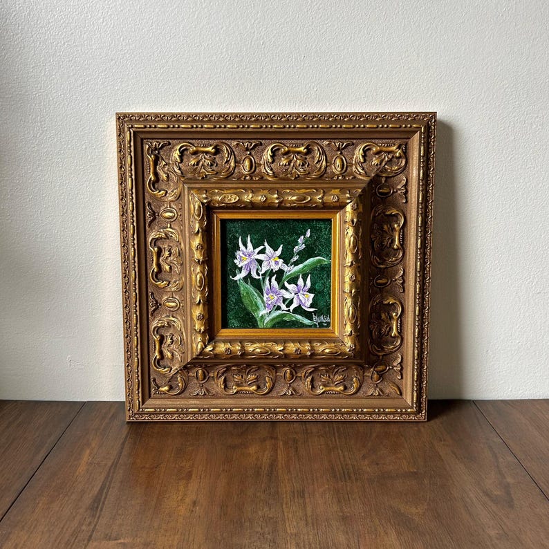May include: Square framed painting featuring white and purple flowers with green leaves against a dark green background. The painting is set within a gold-colored, ornate frame, adding a touch of elegance to the floral artwork.