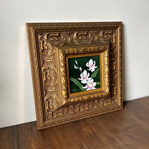 May include: A framed floral painting featuring white orchids with pink centers against a dark green background. The painting is set within a detailed, ornate gold-colored frame. The artwork is displayed on a wooden surface.