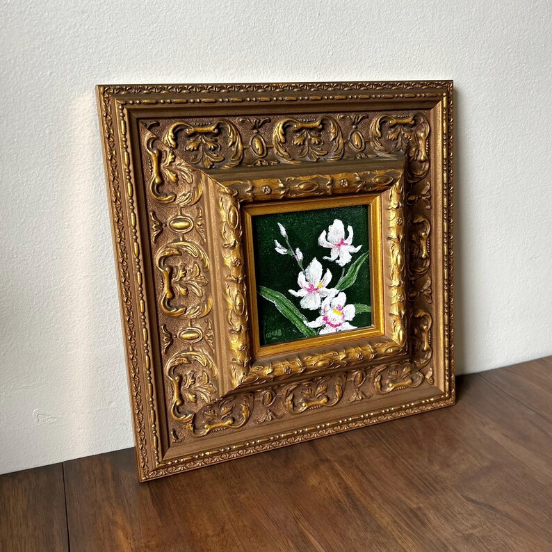 May include: A framed floral painting featuring white orchids with pink centers against a dark green background. The painting is set within a detailed, ornate gold-colored frame. The artwork is displayed on a wooden surface.