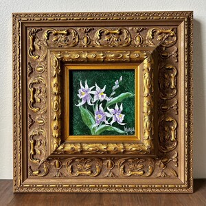 May include: A framed floral painting featuring white and purple orchids with green leaves against a dark green background. The artwork is encased in a detailed, ornate gold-colored frame, adding a touch of elegance.