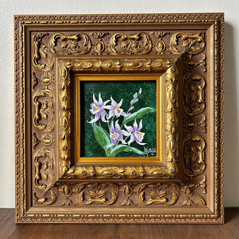 May include: A framed floral painting featuring white and purple orchids with green leaves against a dark green background. The artwork is encased in a detailed, ornate gold-colored frame, adding a touch of elegance.