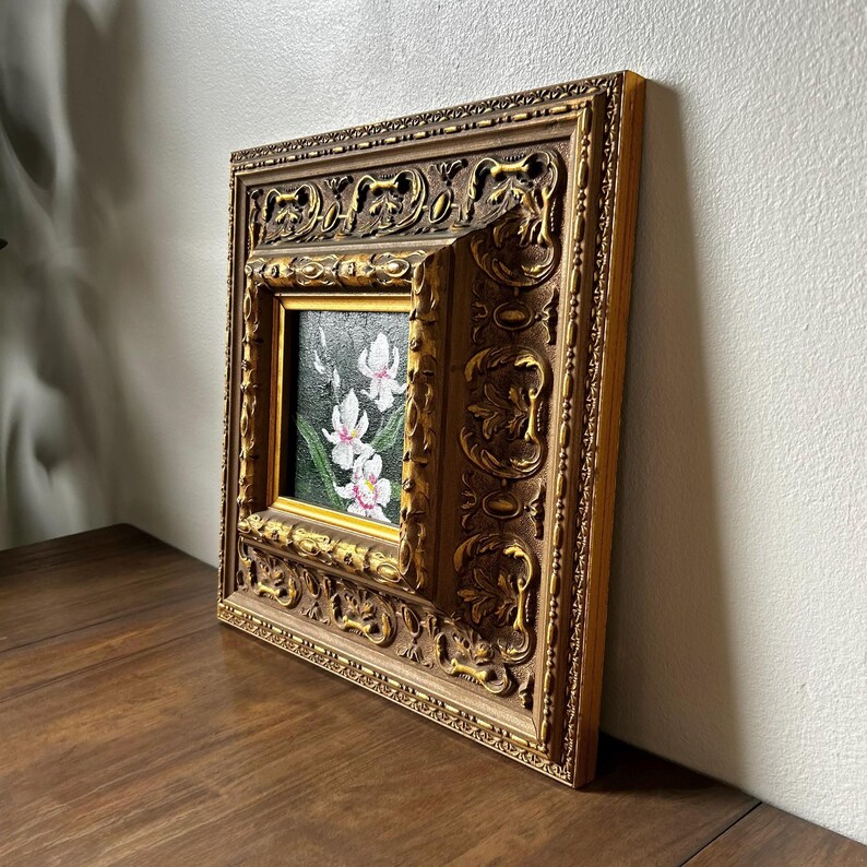 May include: A small, square painting of white and pink orchids in a gold ornate frame. The frame has detailed carvings and a warm, antique gold finish. The painting is set on a dark wooden surface.