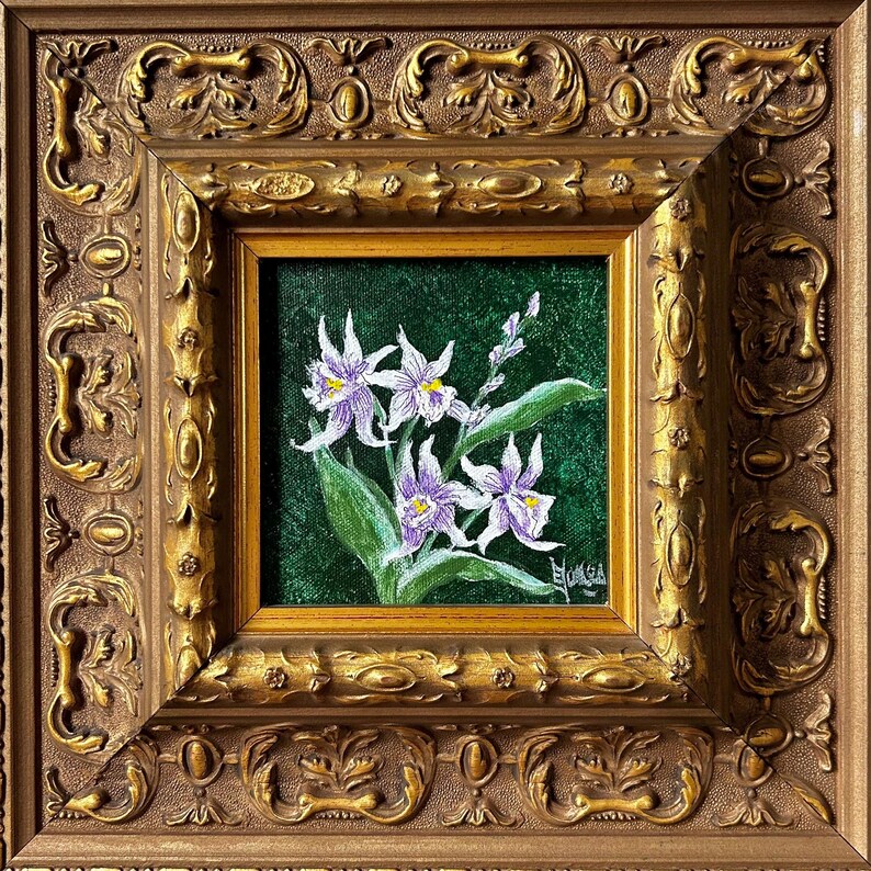 May include: A framed floral painting featuring white and purple orchids with green leaves against a dark green background. The painting is in a gold frame with ornate detailing, encased in a larger, more elaborate gold frame.