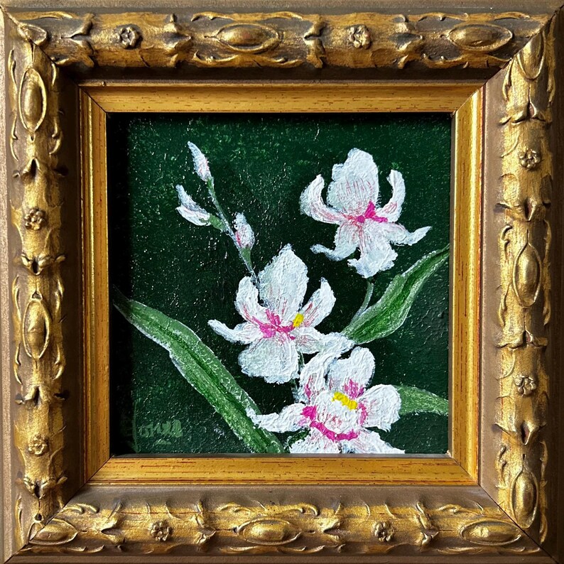 May include: A framed floral painting featuring white orchids with pink and yellow accents against a dark green background. The artwork is enclosed in a gold-colored, ornate frame, adding a touch of elegance to the botanical theme.