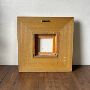 May include: Back of a square, wooden framed painting. The frame is a light brown color with a square opening in the center. A metal sawtooth hanger is attached to the top of the frame.
