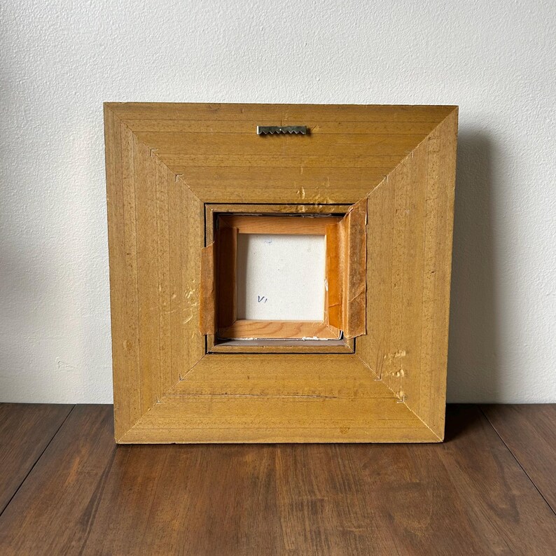 May include: Back of a square, wooden framed painting. The frame is a light brown color with a square opening in the center. A metal sawtooth hanger is attached to the top of the frame.