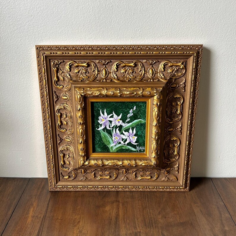 May include: A framed floral painting featuring white and purple flowers with green leaves against a dark green background. The painting is set within a gold-colored, ornate, square frame.