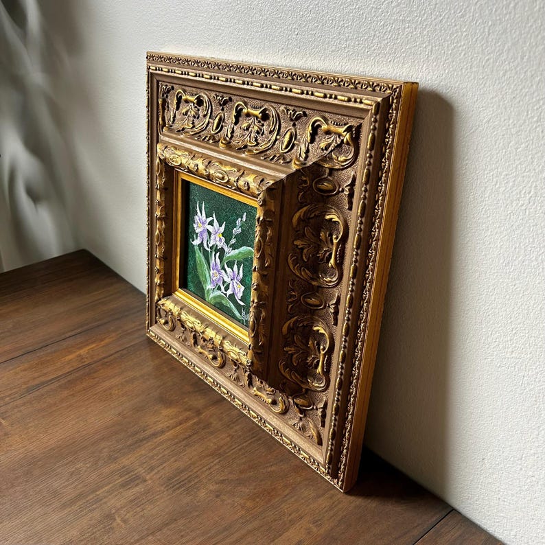 May include: A framed floral painting featuring white and purple flowers with green leaves against a dark green background. The ornate gold frame has detailed carvings. The artwork is displayed on a wooden surface.