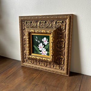 May include: A framed floral painting featuring white orchids with pink accents against a dark green background. The painting is set within a gold-colored, ornate, square frame, adding a touch of elegance to the artwork.