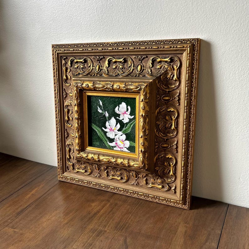 May include: A framed floral painting featuring white orchids with pink accents against a dark green background. The painting is set within a gold-colored, ornate, square frame, adding a touch of elegance to the artwork.