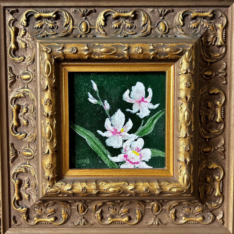 May include: A framed painting of white orchids with pink accents and yellow centers against a dark green background. The painting is in a gold-colored ornate frame.