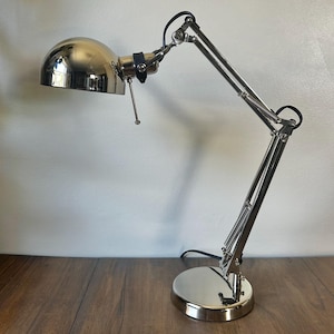 May include: A polished chrome desk lamp featuring a dome-shaped shade, an adjustable articulated arm with visible tension springs, and a round base. The lamp includes a small black leather strap detail near the head and a thin metal pull switch.