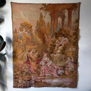 May include: A rectangular wall tapestry featuring a pastoral scene in shades of gold, brown, and muted pink. The design depicts figures in period clothing near a stone fountain, a sphinx statue, and classical columns amidst lush foliage.