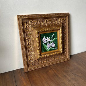 May include: A framed floral painting featuring white and purple flowers with green leaves against a dark green background. The painting is set within a smaller gold frame, which is then set within a larger, ornate gold frame.