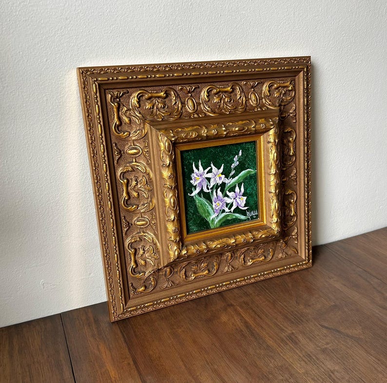 May include: A framed floral painting featuring white and purple flowers with green leaves against a dark green background. The painting is set within a smaller gold frame, which is then set within a larger, ornate gold frame.