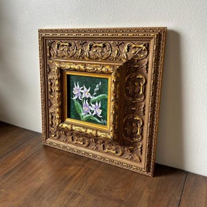 May include: A framed floral painting featuring white and purple flowers with green leaves against a dark green background. The painting is set within a gold-colored, ornate, square frame.