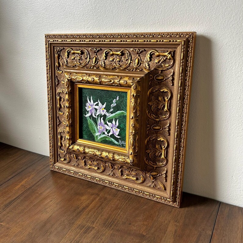 May include: A framed floral painting featuring white and purple flowers with green leaves against a dark green background. The painting is set within a gold-colored, ornate, square frame.