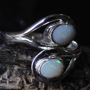 Op de afbeelding: Een zilveren ring met twee ovale opalen. De opalen zijn melkachtig wit met iriserende flitsen. De ring heeft een vloeiend, open ontwerp, met de opalen gezet in bezel-zettingen.
