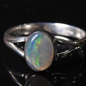 Op de afbeelding: Een zilveren ring met een ovale opaal edelsteen. De opaal toont iriserende kleuren van groen, blauw en geel. De ringband heeft een gespleten ontwerp, wat een uniek detail aan de sieraden toevoegt.