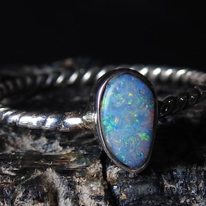 Op de afbeelding: Zilveren ring met een peervormige opaalsteen. De opaal toont een levendig kleurenspel, met blauw, groen en andere tinten. De band heeft een gedraaid touwontwerp, wat textuur aan de ring toevoegt.