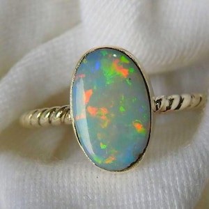 Op de afbeelding: Een gouden ring met een ovale opaal edelsteen. De opaal vertoont een levendig kleurenspel met flitsen van groen, oranje en blauw. De ringband heeft een gedraaid touwontwerp, wat een klassieke touch aan de sieraden toevoegt.