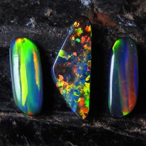 May include: Three polished opal gemstones. The opals display a vibrant array of colours, including green, blue, orange, and yellow. The gemstones are set against a dark, textured background, highlighting their iridescent qualities.