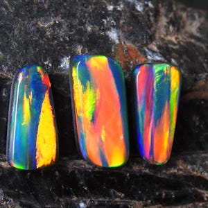 Könnte beinhalten: Drei schillernde schwarze Opale mit einer Vielzahl von Farben, darunter Blau, Grün, Gelb, Orange und Rot. Die Opale sind auf einem dunkelgrauen Hintergrund platziert.
