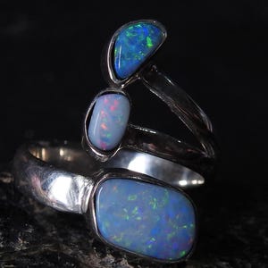 Op de afbeelding: Een zilveren ring met drie opaalstenen. De opalen zijn blauw met groene en roze flitsen. De ring heeft een uniek, open ontwerp met de stenen in verschillende hoeken geplaatst.