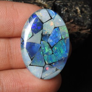 Könnte beinhalten: Ovale Edelstein mit Mosaikdesign. Der Edelstein zeigt eine Kombination aus blau, weiß und grün schillernden Teilen, die ein einzigartiges Muster bilden. Der polierte Stein hat ein glänzendes Finish.