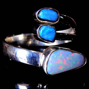 Op de afbeelding: Zilveren ring met drie opalen. Twee ovale, blauwe opalen zijn bovenop geplaatst, en een grotere, driehoekige opaal met blauwe, roze en rode tinten aan de basis. De ring staat op een zwarte achtergrond.