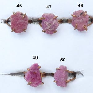 Puede incluir: Cinco anillos con piedras preciosas rosas en bruto engastadas en metal color cobre. Las piedras tienen una textura natural y rugosa. Los anillos se muestran sobre una superficie blanca. Los anillos están numerados 46, 47, 48, 49 y 50.