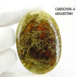 May include: An oval-shaped cabochon gemstone featuring a polished surface with swirling patterns of olive green, brown, and amber tones. The stone is labelled CABOCHON-A 68X48X7MM, indicating its dimensions in millimetres as 68 by 48 by 7 mm.