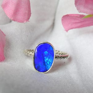 Op de afbeelding: Een zilveren ring met een grote, ovaalvormige blauwe opaal edelsteen. De opaal heeft een levendige, iriserende kwaliteit. De band van de ring heeft een gedraaid touwontwerp. De ring is geplaatst tegen een witte achtergrond met roze bloemblaadjes.
