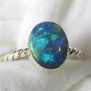Op de afbeelding: Zilveren ring met een ovale opaal. De opaal heeft een levendig kleurenspel van blauw en groen. De band heeft een gedraaid ontwerp. De ring staat tegen een witte achtergrond.