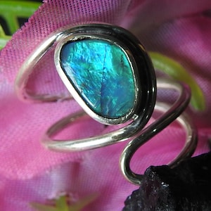 Op de afbeelding: Zilveren ring met een ovale, iriserende edelsteen in blauw- en groentinten. De ring heeft een uniek, wervelend bandontwerp. De edelsteen is geplaatst tegen een roze en groene achtergrond. Een opvallend sieraad.