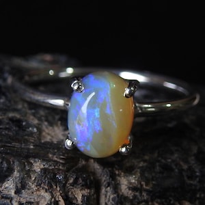 Op de afbeelding: Een zilveren ring met een ovale opaal edelsteen. De opaal toont blauwe, gele en oranje tinten. De edelsteen wordt vastgehouden door vier zilveren pootjes. De ring staat tegen een donkere achtergrond.
