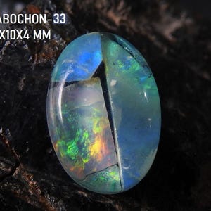 Könnte beinhalten: Ein ovaler Cabochon-Edelstein mit schillernden Farben in Blau, Grün und Gelb. Der Stein ist in der Mitte gespalten. Der Text oben lautet "CABOCHON-33" und "14x10x4 MM". Der Edelstein befindet sich auf einem dunklen, strukturierten Hintergrund.
