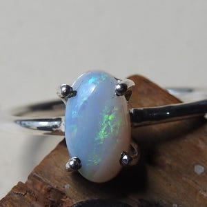 Op de afbeelding: Een zilveren ring met een ovale opaal edelsteen. De opaal heeft een lichtblauwe basiskleur met groene flitsen en wordt vastgehouden door vier zilveren klauwen. De ringband is van gepolijst zilver.