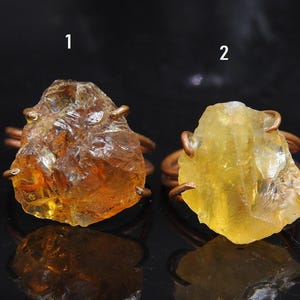 May include: Two raw gemstone rings. Ring 1 has a rough, amber-coloured stone. Ring 2 displays a yellow-hued stone. Both stones are set on copper-coloured wire ring bands. The rings are displayed on a reflective black surface.